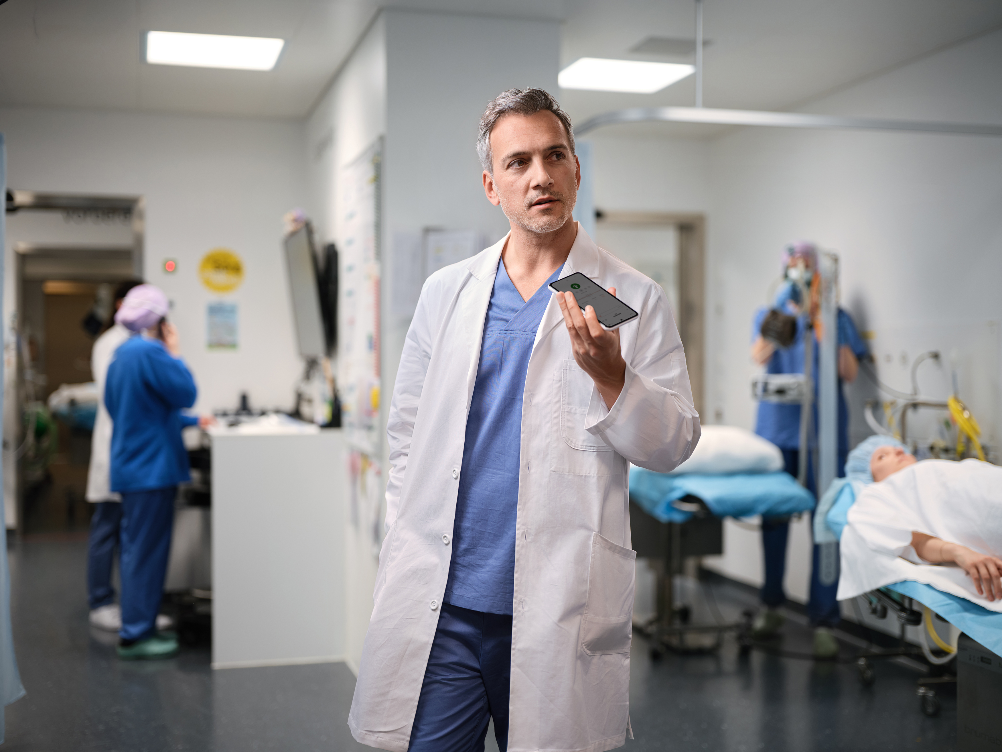 Un médecin se tient dans la salle d'opération et dicte dans son téléphone portable. À l'arrière-plan, on aperçoit du personnel médical spécialisé en train de préparer l'opération.