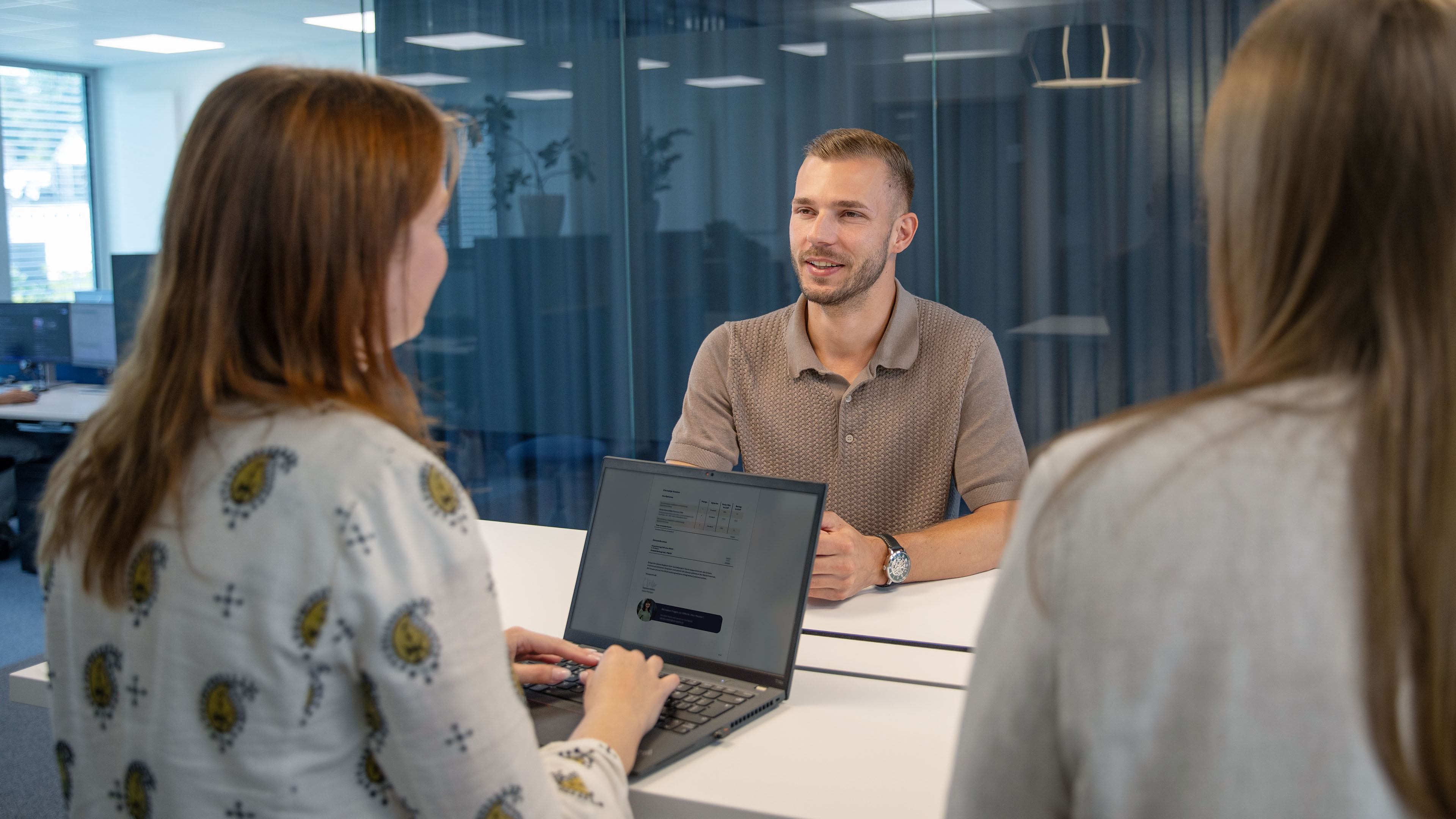 Drei Mitarbeitende sitzen an einem Tisch und führen ein Gespräch, eine Person arbeitet dabei am Laptop