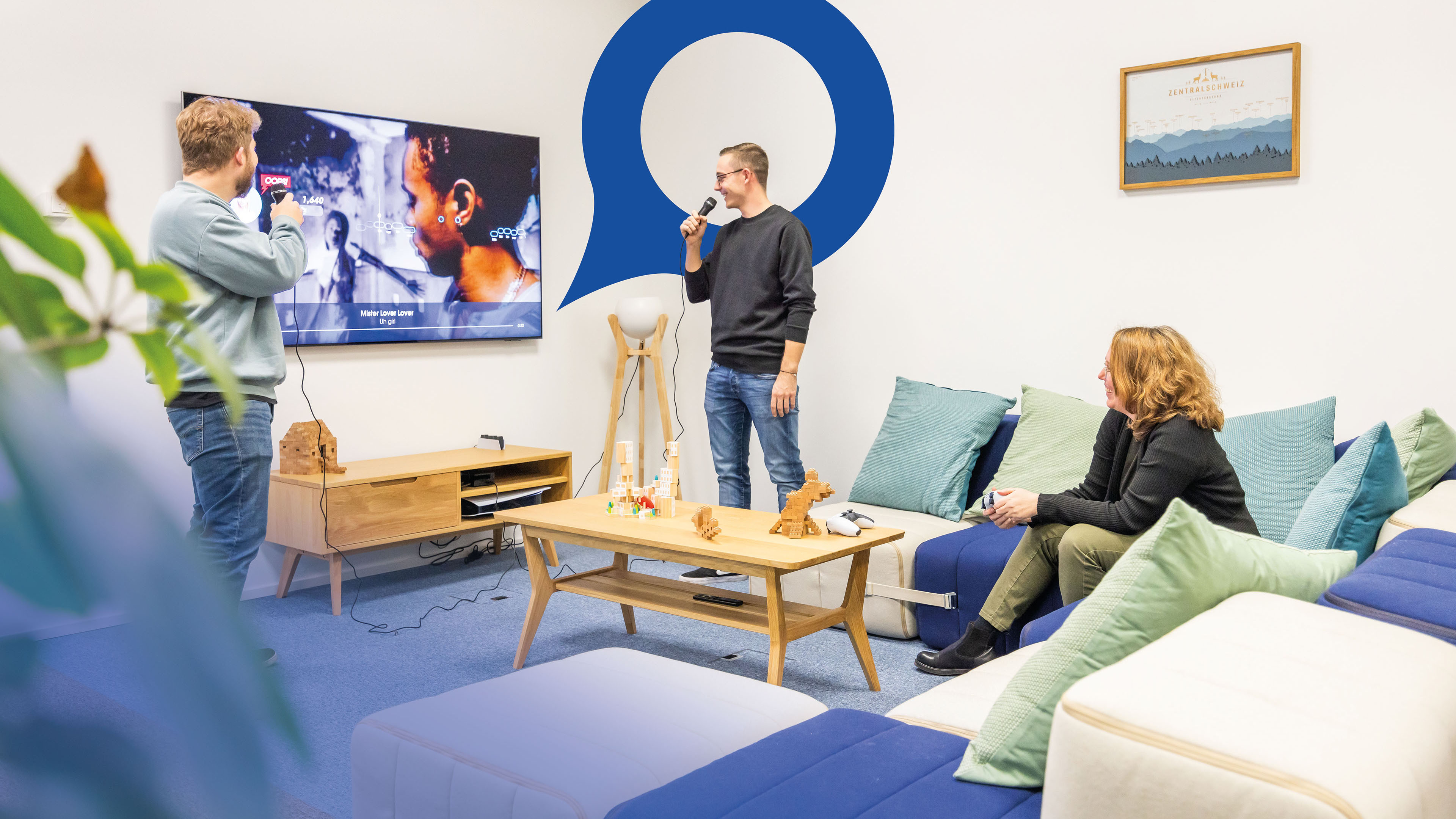 Two employees are standing in the break room of the Voicepoint office in Wetzikon singing karaoke. The blue Voicepoint speech bubble is visible in the background.