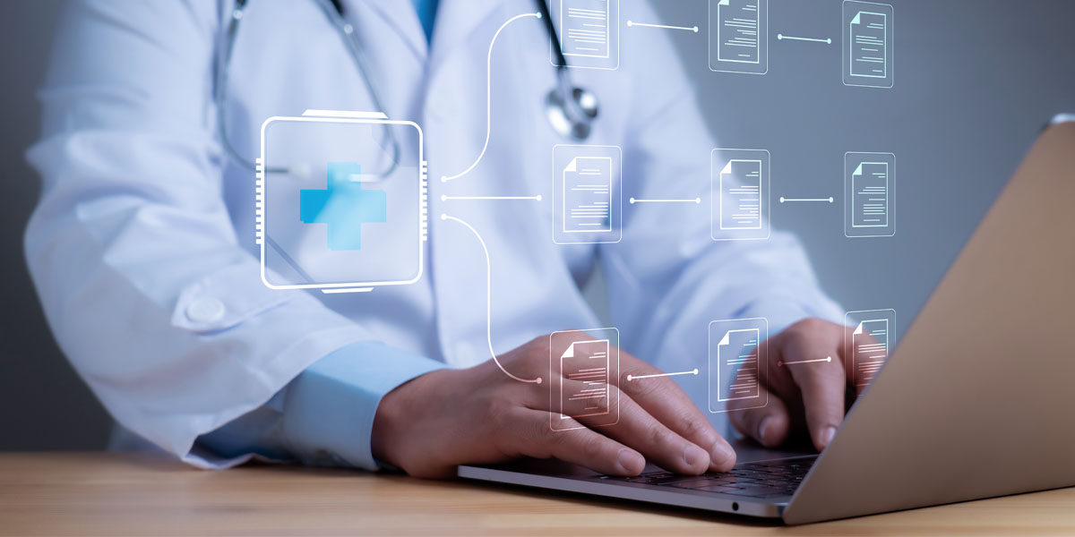 Arzt im weissen Kittel sitzt am Schreibtisch und dokumentiert mit PC eine elektronische Patientenakte.