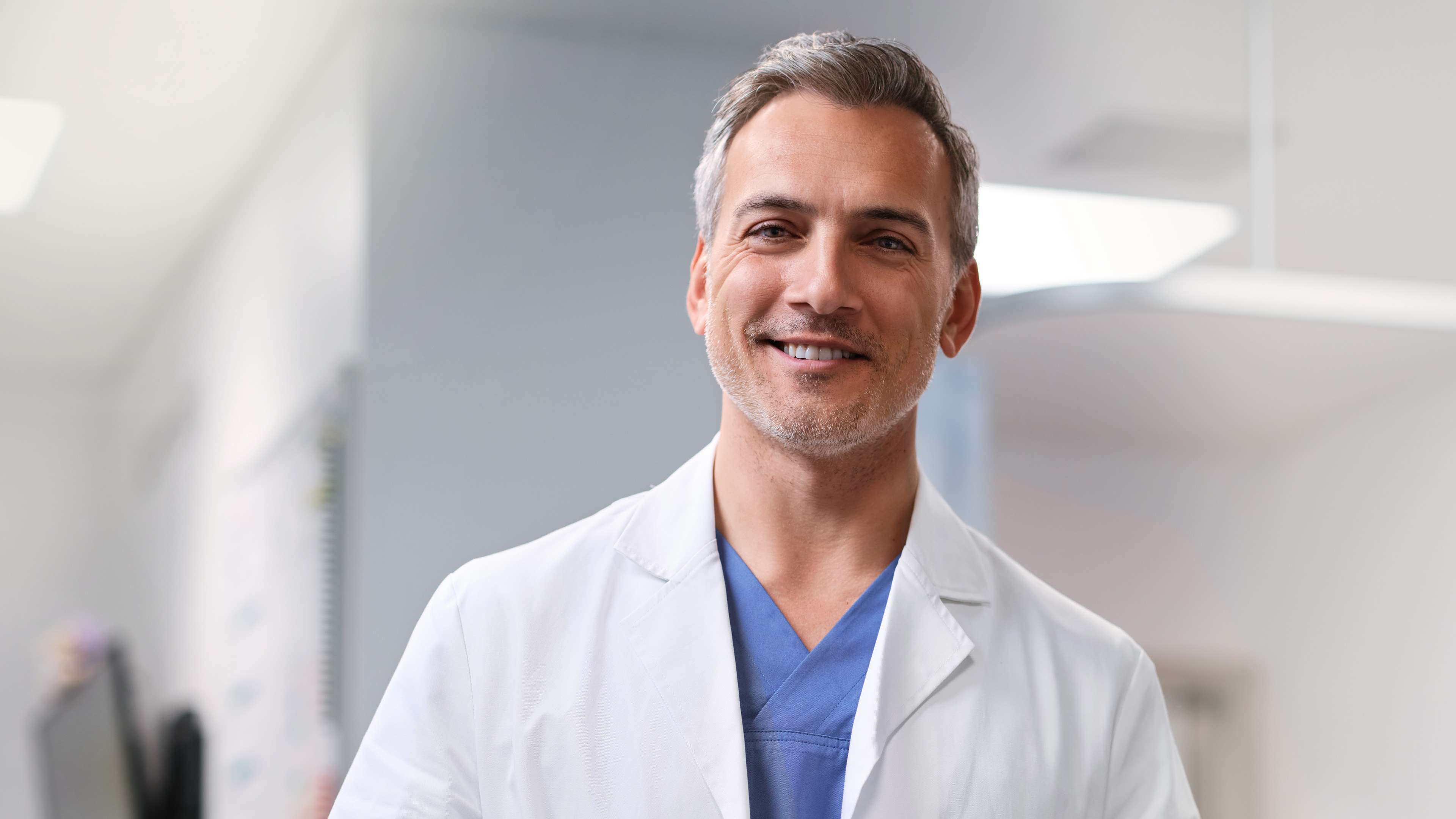 A doctor in a white coat stands in a hospital corridor and smiles at the camera.