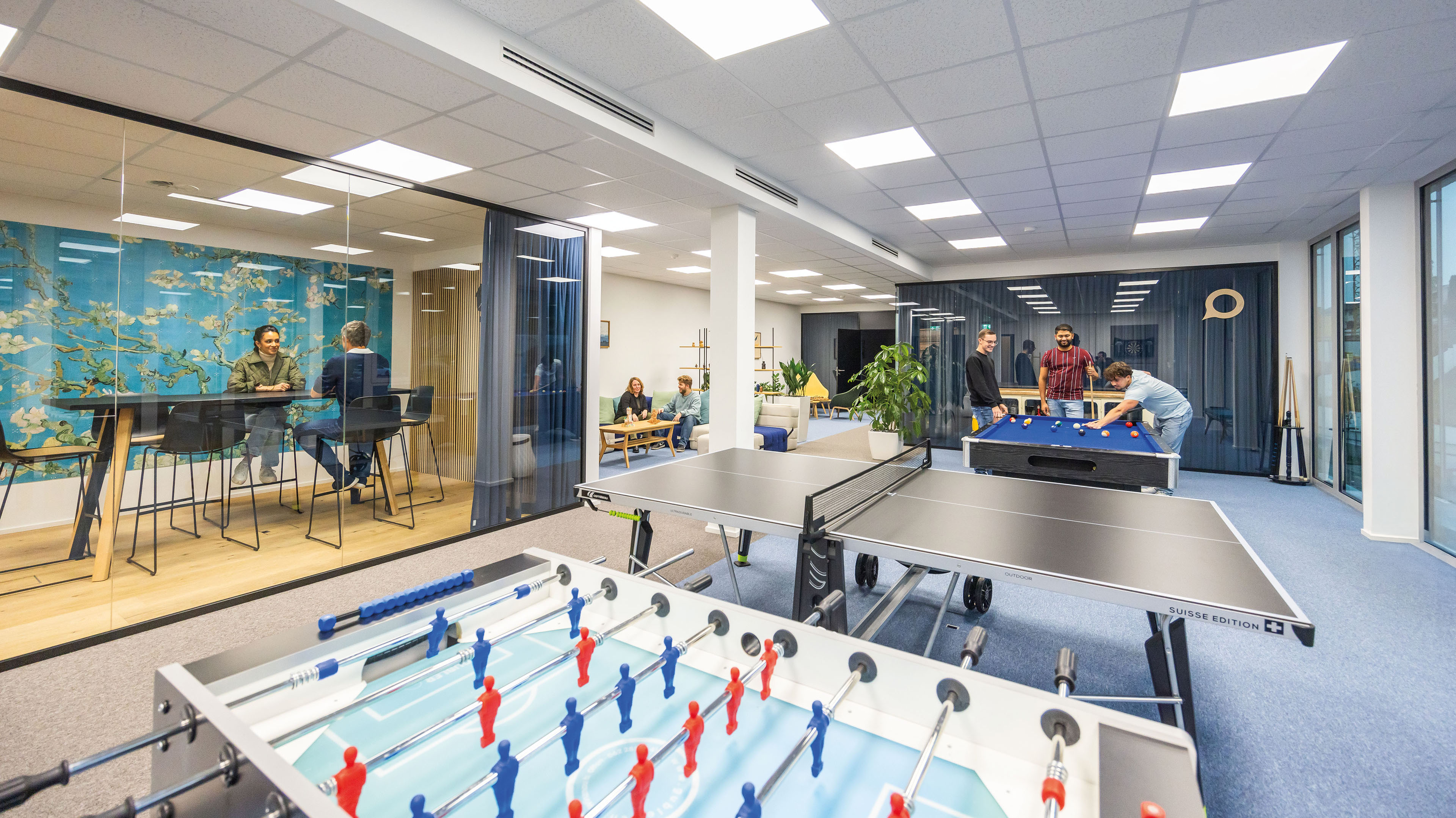 Mitarbeitende spielen Billard und sitzen zusammen im Aufenthaltsbereich, vorne sind ein Pingpongtisch und Tischfussball zu sehen.