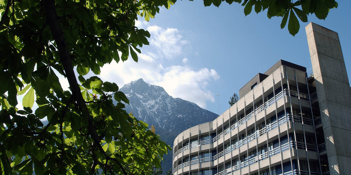 Gebäude des Hôpital du Valais mit Blick auf die Alpen, eingerahmt von grünen Baumästen im Vordergrund.