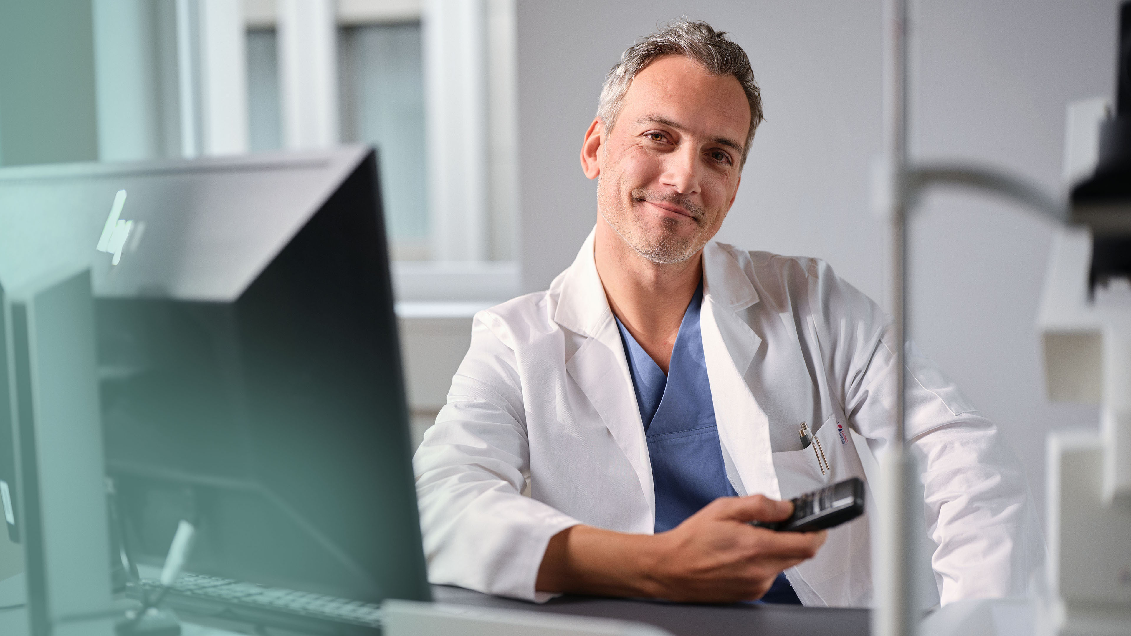 Un médecin en blouse blanche est assis à son poste de travail devant un ordinateur Windows et dicte avec Dragon Medical One.