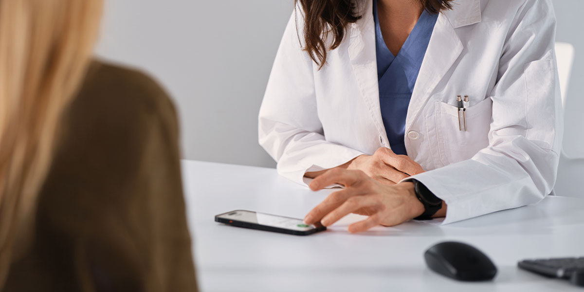 Ärztin sitzt in einem Behandlungszimmer der Patientin gegenüber und nimmt die Sprechstunde mit einem Smartphone auf.